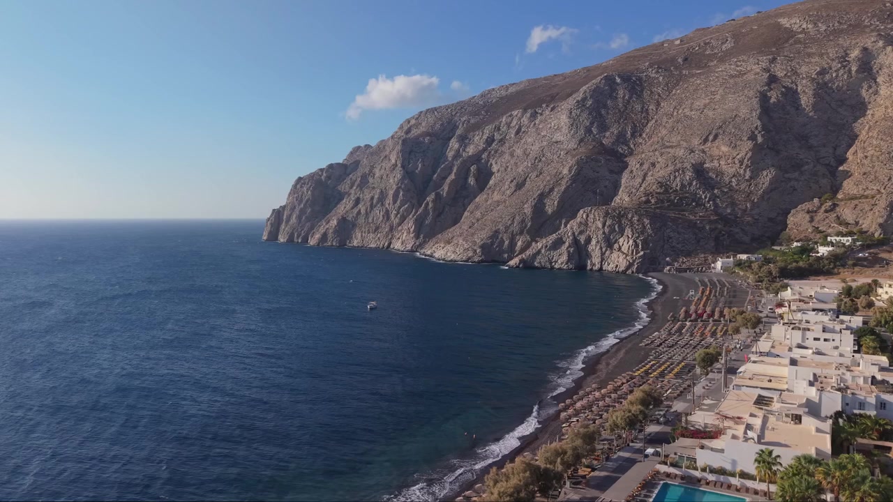 Drohnenaufnahme, die den Kamari Beach auf Santorin zusammen mit der umliegenden Landschaft enthüllt, mit Klippen, Blicken auf die Caldera und dem tiefblauen Ägäischen Meer.