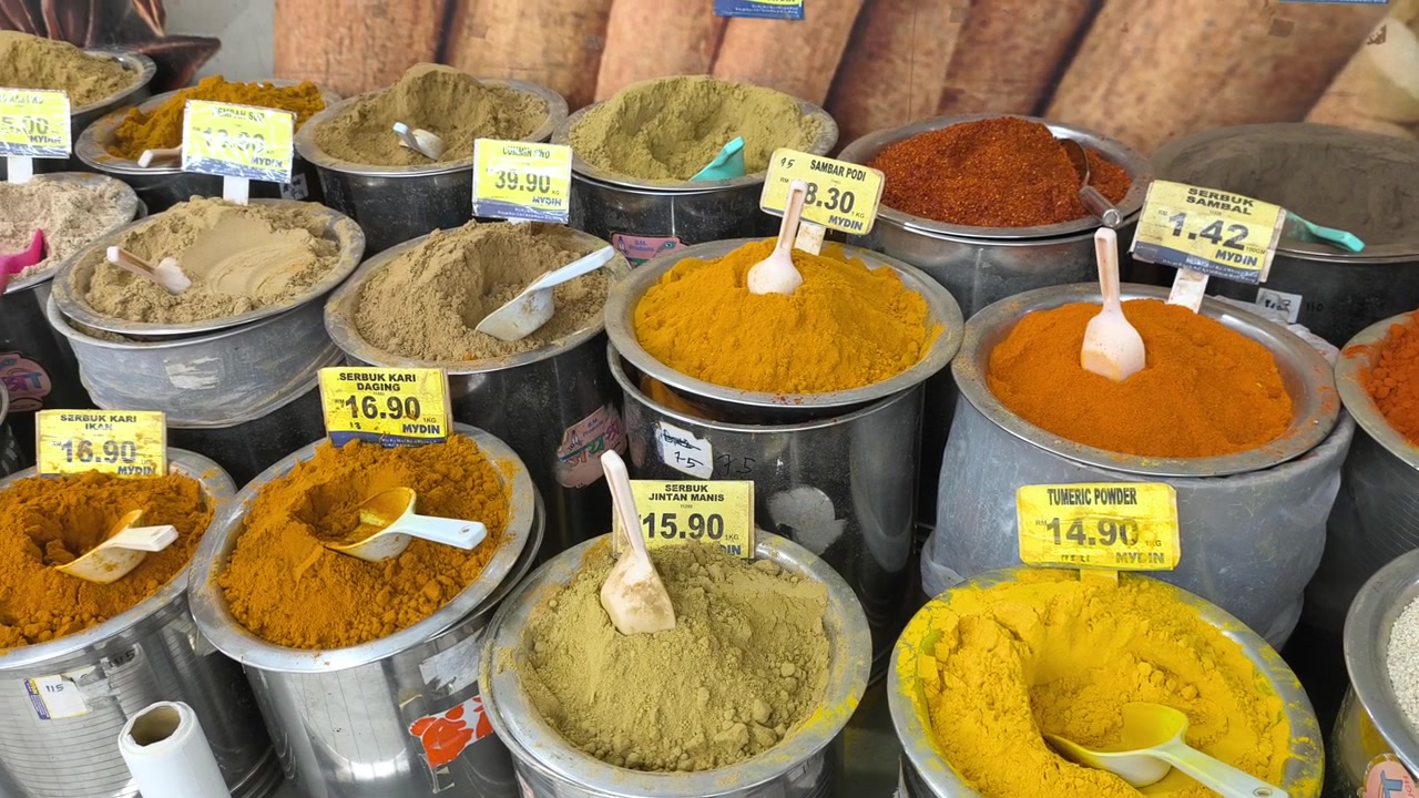 A variety of colorful spices are neatly displayed for sale in clean stainless steel jars. Each container features a clear price tag. The organized market setup looks hygienic and appealing.