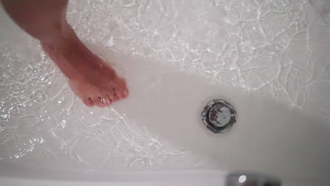 woman foot in clear water in bathtub closeup. bare lady leg and dripping sprays in shower cabin. woman stands in warm liquid in home washroom