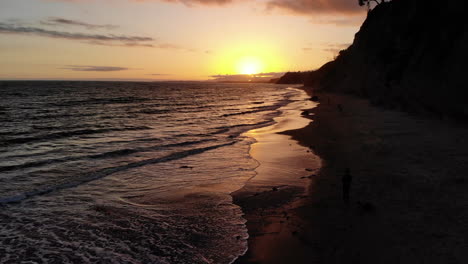 disparo aéreo de un dron volando bajo sobre la playa de arena con vibrantes reflejos del atardecer en las olas del océano frente a la costa de santa bárbara, california