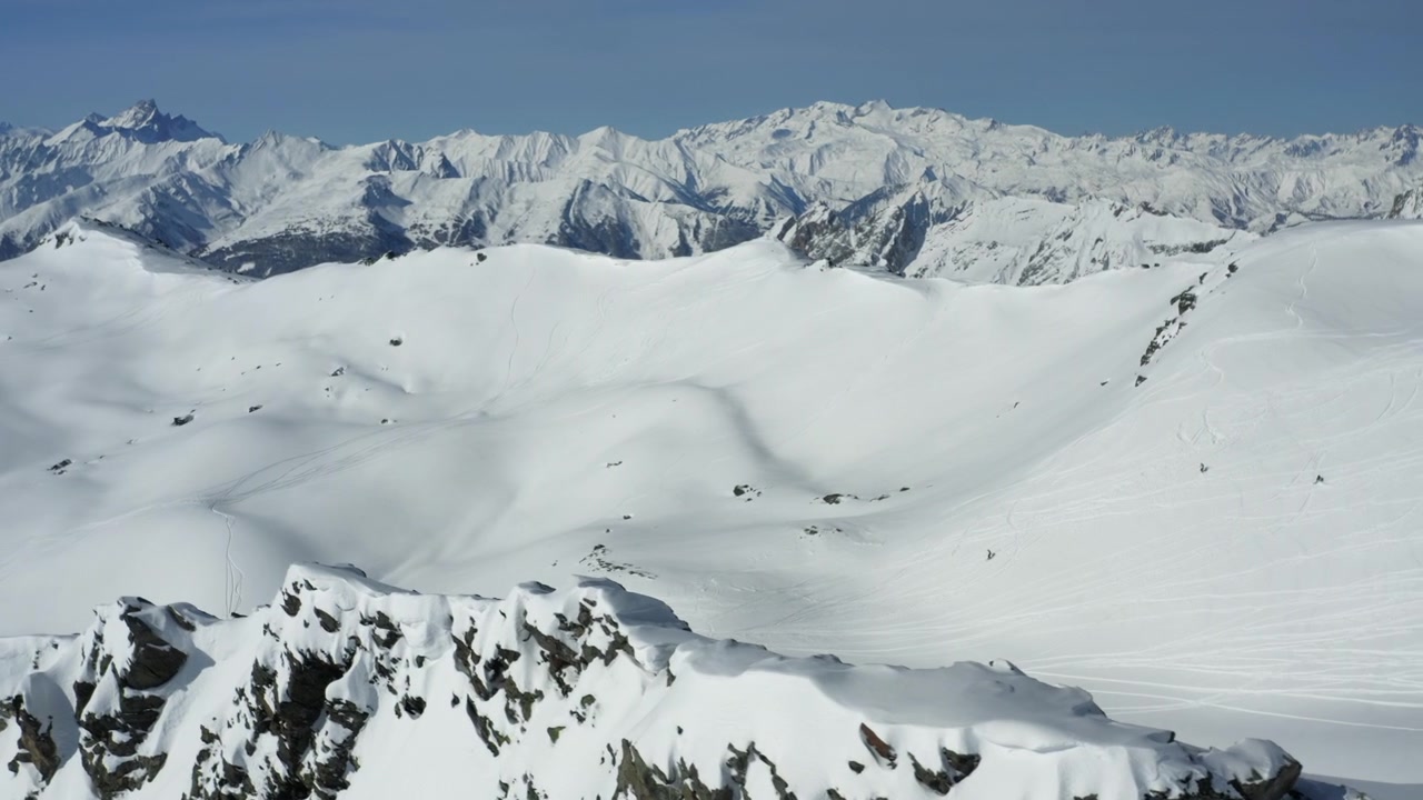 Drone flies along snowy mountain ridge exposing a vast valley with a group of skiers freeriding fresh snow under clear blue sky. Pristine winter landscape, distant mountain ridge. Parallax, blocking.
