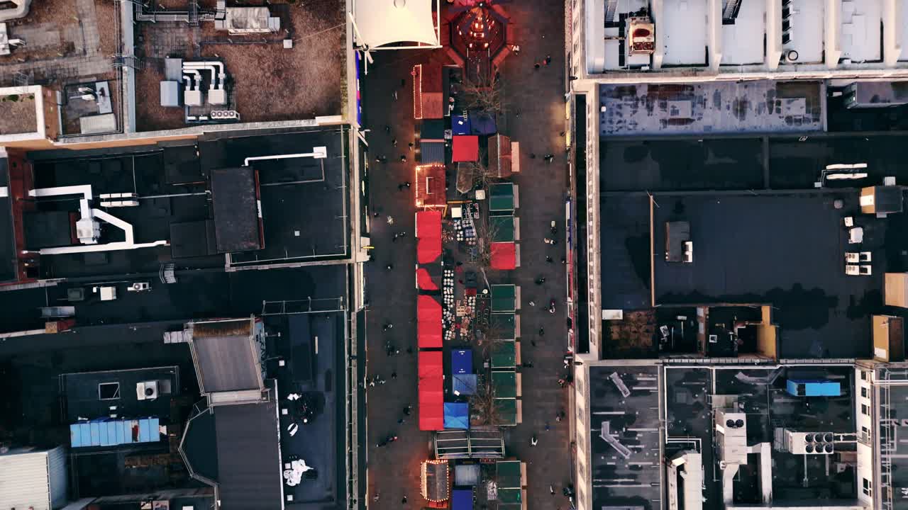 Landscape drone shot rising into a bird’s-eye view over Southampton’s Christmas Market in winter at sunset, revealing glowing festive stalls, crowds exploring the square, and warm evening light