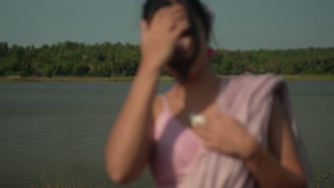 Woman-in-pink-saree-by-the-lake-at-golden-hour,-touching-necklace,-thoughtful-expression