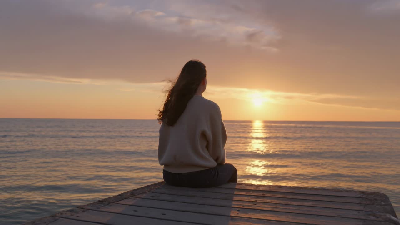 Sol hundiéndose en el horizonte, mujer sentada en el muelle frente al océano, el cabello elevándose, el cielo cambiando evocando calma.