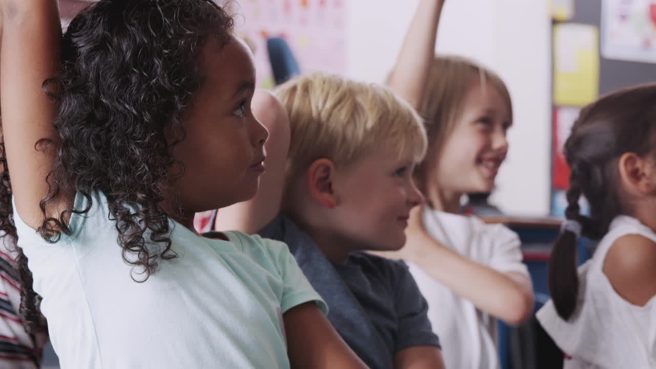 Group Of Elementary School Pupils Sitting On Floor Raising Hands To Answer Question