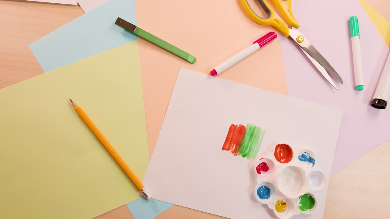 Overhead view of hand painting colorful swatches on paper, surrounded by art supplies, bright lighting