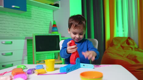 adorable smiling caucasian toddler in blue sweater sits at desk. cute child uses molding machine to make plasticine figures.