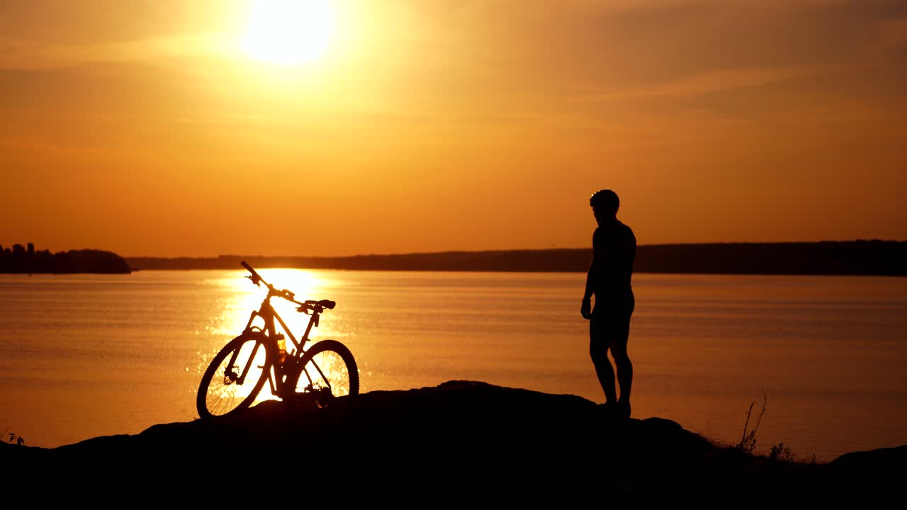 Sports bike and bicyclist at sunset near the river. Amazing view of golden path over the water surface on the bike and a silhouette of a man outdoors.