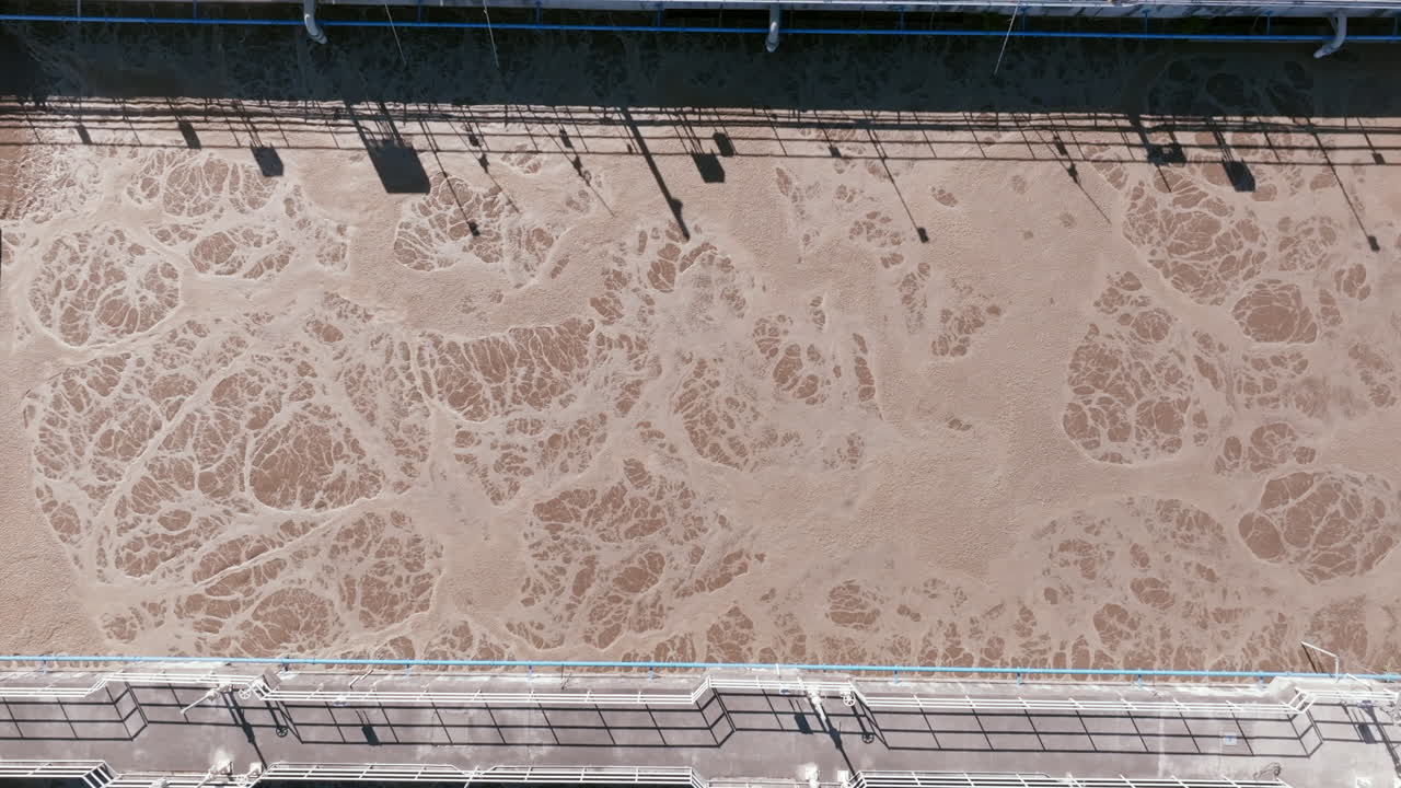 A zoomed-in aerial shot of one aeration basin at the Spokane facility shows swirling tan foam textures with a pedestrian walkway and railing overhead