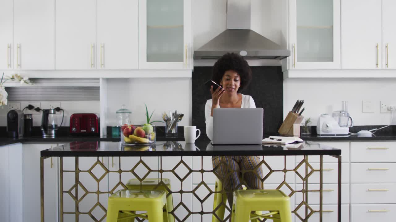 African american woman talking on smartphone and using laptop in the kitchen while working from home