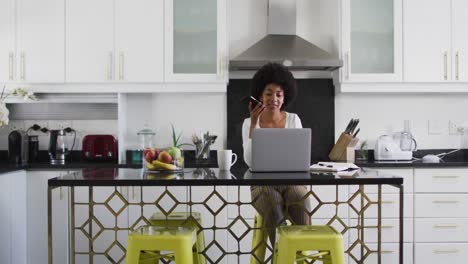 african american woman talking on smartphone and using laptop in the kitchen while working from home