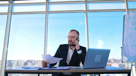portrait of entrepreneur at workplace. confident businessman in glasses talking the phone while sitting in front of a laptop in office.