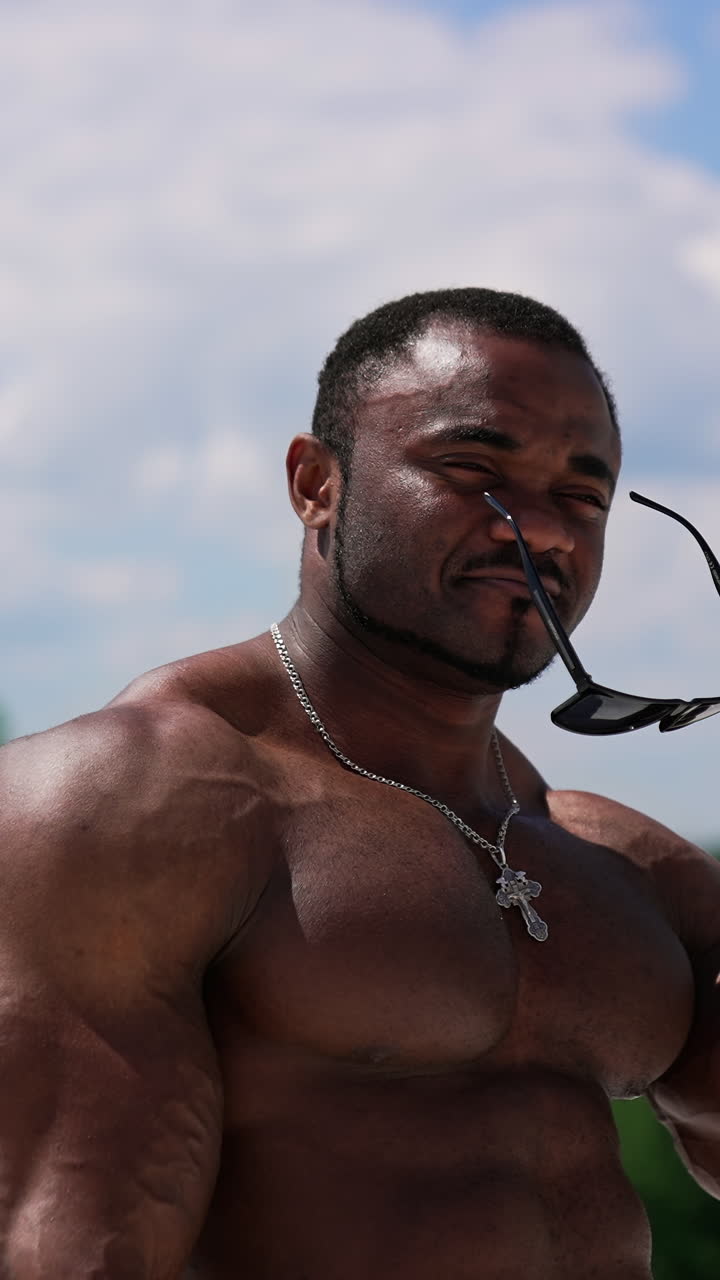 Waist up portrait view of the strong stylish man wearing glasses while posing on clear blue sky background. Shirtless bodybuilder showing his muscles. Vertical video