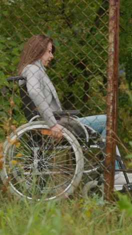 joven en silla de ruedas descansa moviéndose en el parque más allá de la vieja valla de malla. restauración de la salud en el complejo turístico. rehabilitación después de lesiones en las piernas vista lateral