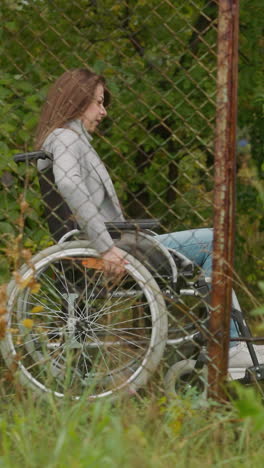 young woman in wheelchair rests moving in park past old mesh fence. health restoration at resort complex. rehabilitation after legs injury side view