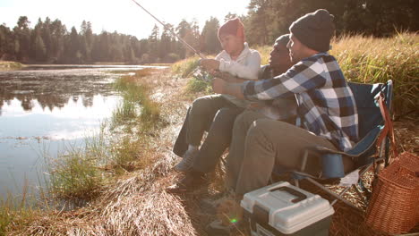 grandfather and father sit teaching boy to fish by a lake