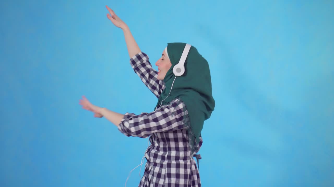 히자브를 입은 젊은 이슬람 여성의 초상화. 헤드폰을 들고 음악을 듣고 파란색 배경에서 춤을 춘다.