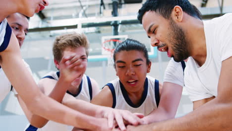 Jugadores-Masculinos-De-Baloncesto-De-Secundaria-Uniendo-Sus-Manos-Sobre-La-Pelota-Durante-La-Charla-En-Equipo-Con-El-Entrenador