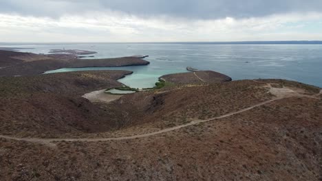 Playa-Balandra-beach-and-surrounding-mountains-near-La-Paz-in-Baja-California-sur-with-clear-blue-waters-and-rugged-terrain,-under-cloudy-skies,-aerial-view