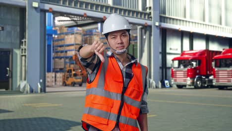 asian male engineer with safety helmet showing thumbs down gesture and shaking his head while standing , outside of logistics distributions warehouse