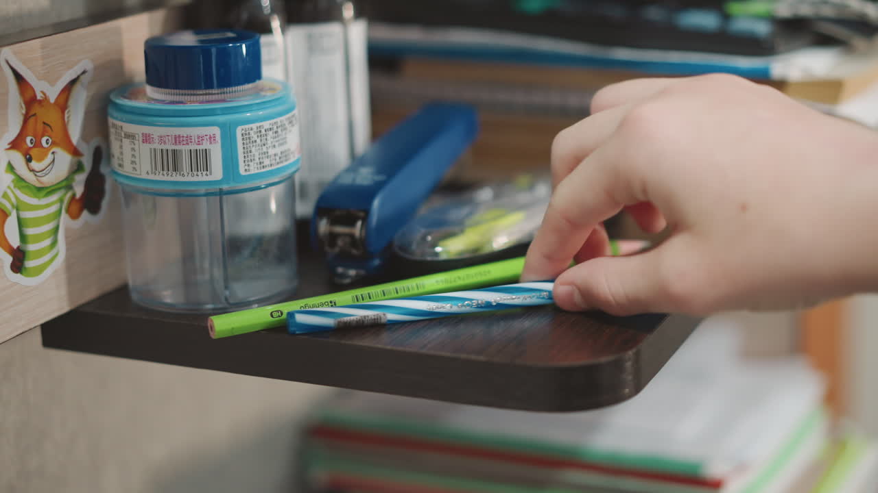 Wooden shelf with pencils jar and stapler, grandson reaches to arrange colorful pens on edge, stacked books in background, soft natural light, cozy study corner, intimate household moment