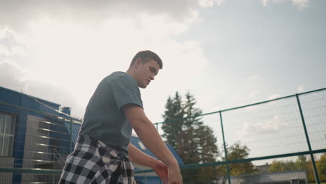 man bouncing volleyball outdoors with his right hand, sunlight creating a silhouette effect around his shoulder and head, with a blurry background of sports arena and trees