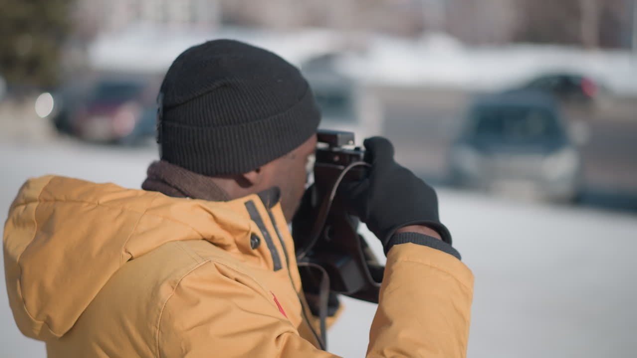 back side view of black man fixing gaze through vintage camera viewfinder while delicately adjusting lens focus dial against snowy urban plaza under bright winter sun