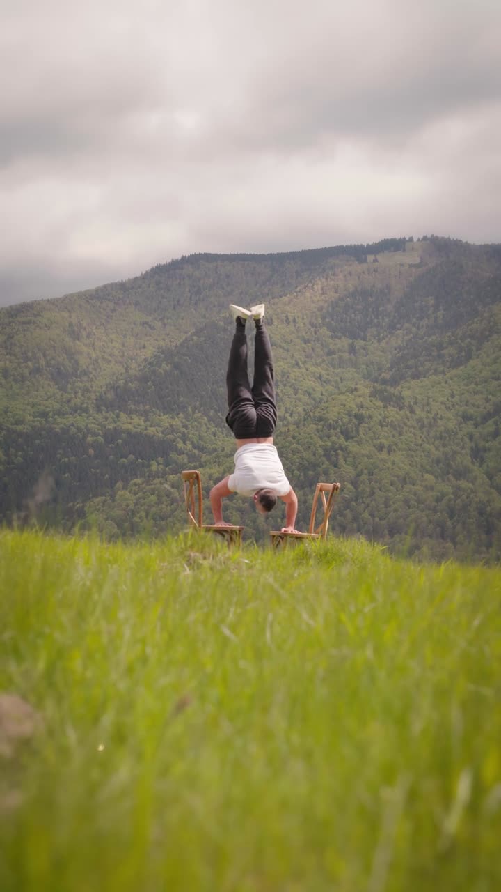 Athlete do handstand balanced on two chairs against a scenic green nature backdrop