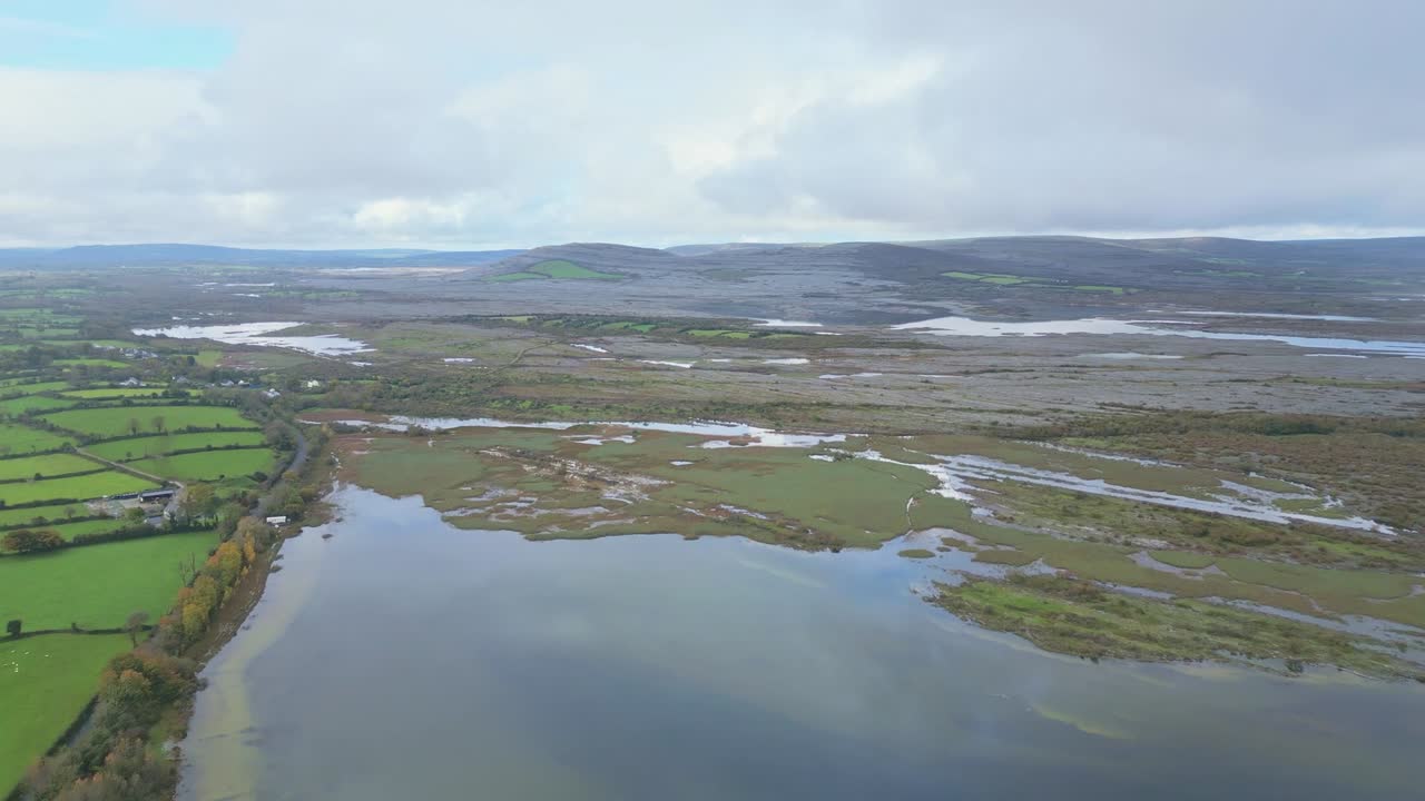 Luftfoto af en afsides irsk kystlinje i Burren-regionen, med grønne marker, kalkstensbakker og reflekterende vand under overskyet himmel.