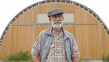 portrait shot of caucasian senior farmer man in hat standing outdoors at a stable, looking at side and turning his face to the camera