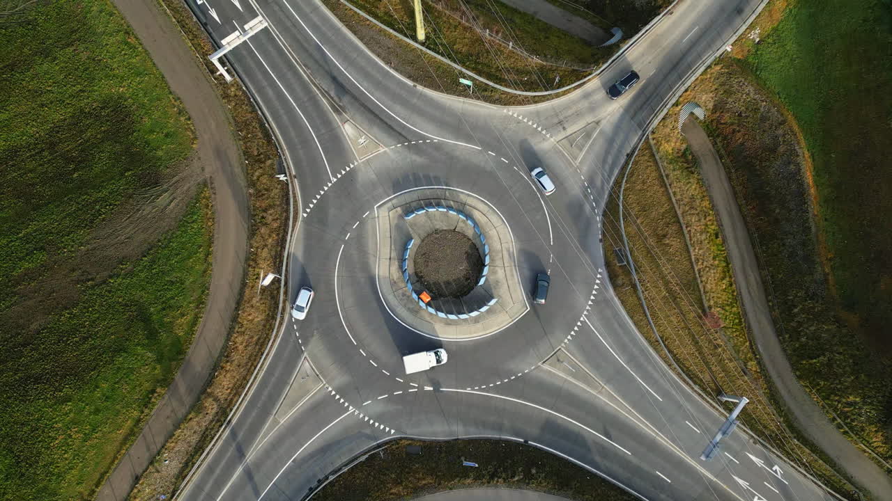 Drone shot looking straight down at a multi-lane roundabout with cars and trucks circulating smoothly, surrounded by rural roads and fields