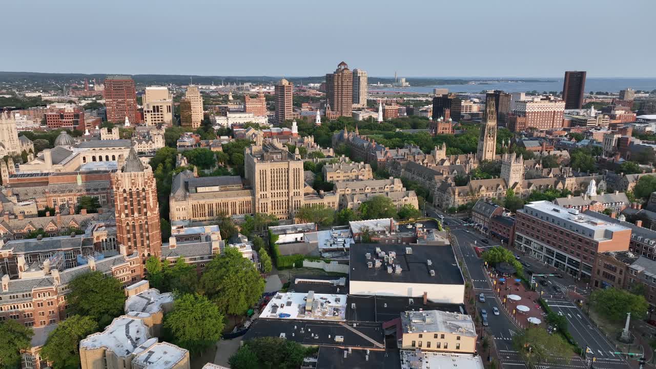 Cars on street of New Haven downtown at sunrise. Historic city with brick houses and buildings. Gothic Yale university campus and harkness tower. Long Island Sound Port in background. Aerial wide shot