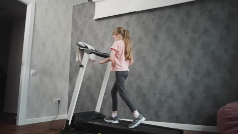 rear view of young girl in patterned leggings walking on treadmill during home workout session long hair tied back hands on treadmill handles grey textured wall and wooden floor visible
