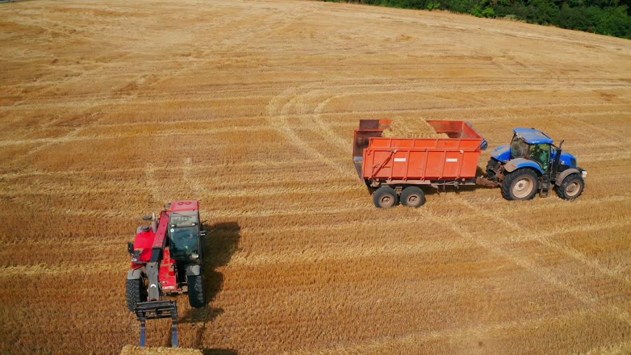 Big tractor machine being loaded with straw. Skid loader picks up two hay bales piled on one another. Aerial view.