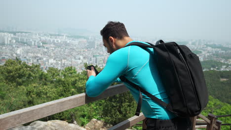 Hombre-De-Senderismo-Con-Mochila-Usando-Teléfono-Móvil-En-Lo-Alto-De-La-Pista-Forestal-De-La-Montaña-Gwanaksan-Con-Vistas-A-La-Ciudad-De-Seúl-En-El-Fondo