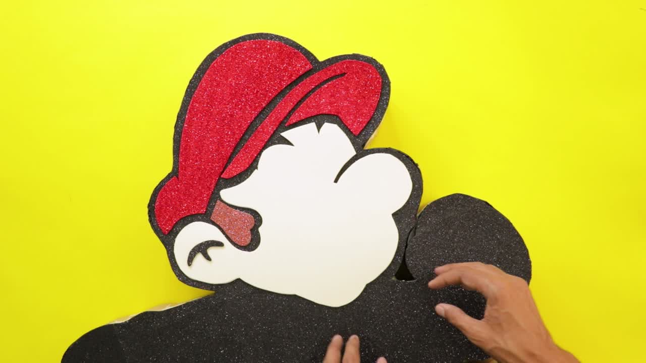 Hands assembling a sparkly foam cutout of a red-capped video game character against a yellow background