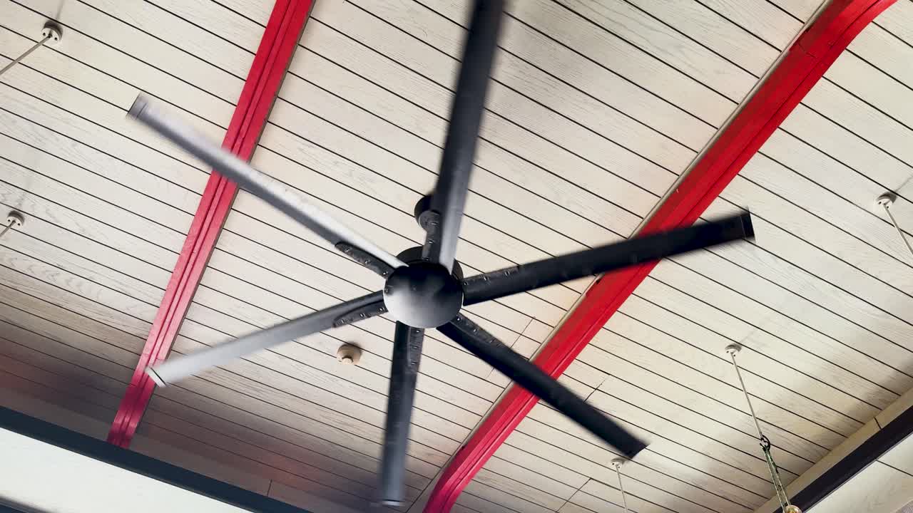 A large ceiling fan rotates steadily against a wooden ceiling with red beams, creating a dynamic visual effect