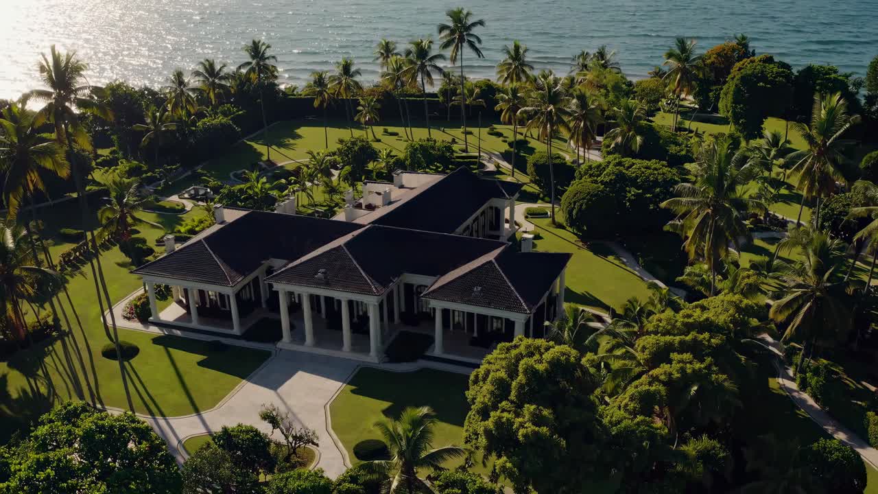 Aerial video view of a luxurious beachfront mansion surrounded by lush greenery and palm trees