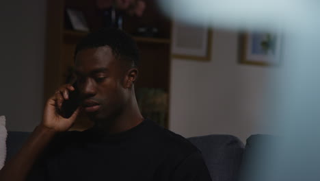 unhappy and depressed young man sitting on sofa at home making call on mobile phone at night