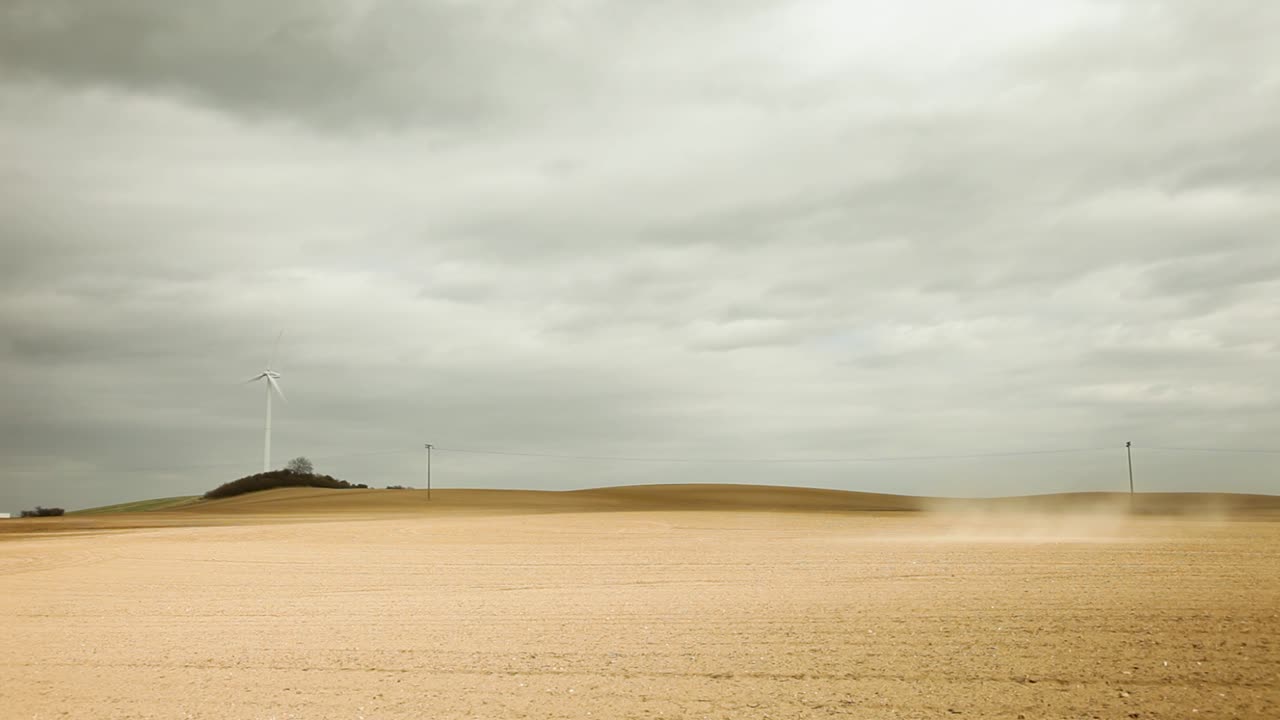 una turbina eólica blanca solitaria se encuentra en un campo estéril de color marrón claro bajo un cielo nublado gris, evocando un estado de ánimo desolado, con una colina, árboles, líneas eléctricas y una tormenta de polvo en la distancia