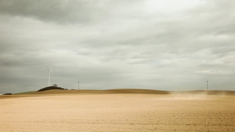 Eine-Einsame-Weiße-Windkraftanlage-Steht-Auf-Einem-Kargen-Hellbraunen-Feld-Unter-Einem-Grau-Bewölkten-Himmel-Und-Ruft-Eine-Trostlose-Stimmung-Hervor,-Mit-Einem-Hügel,-Bäumen,-Stromleitungen-Und-Einem-Staubsturm-In-Der-Ferne