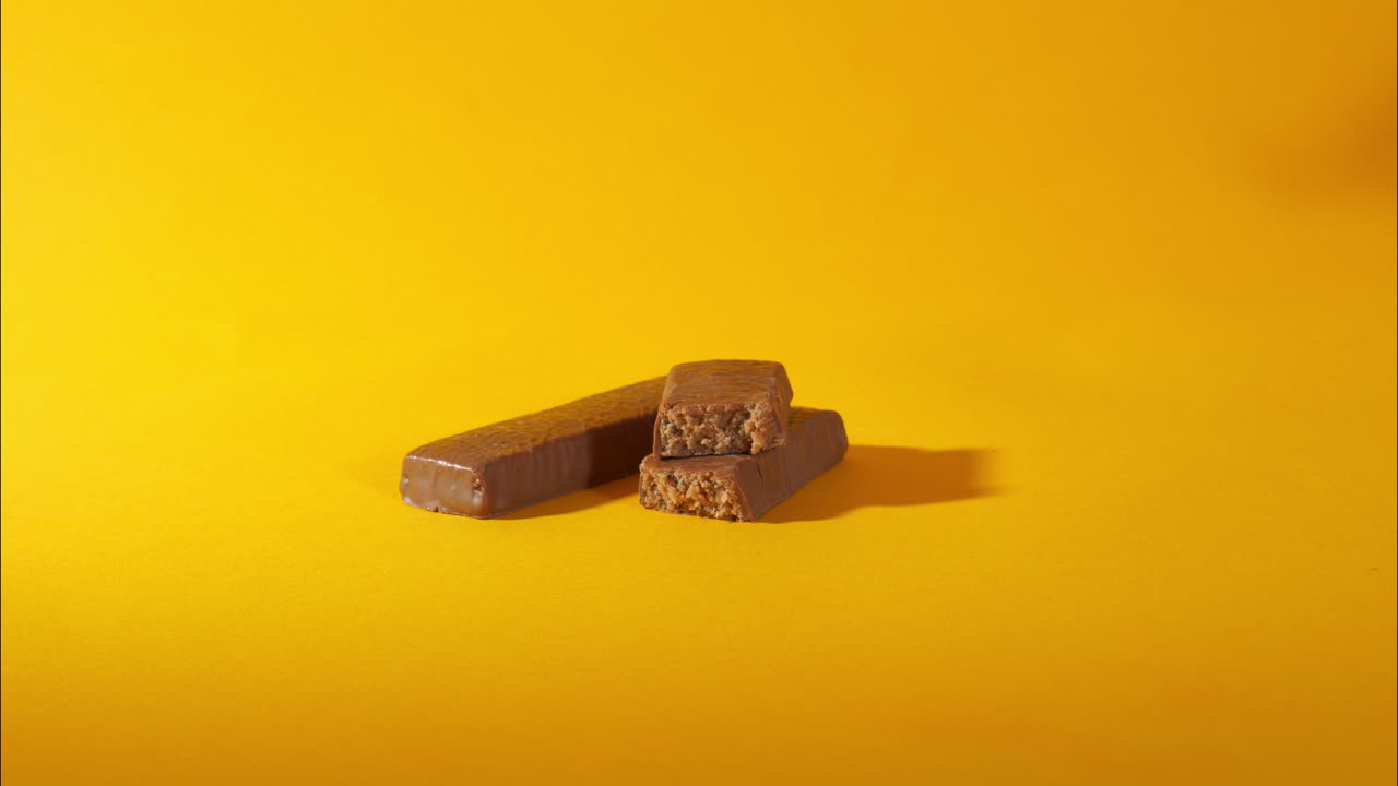 A hand grabs a stacked vegan protein chocolate bar with a bite showing the textured interior, displayed on a vibrant yellow surface—highlighting ingredients like soy, pea, rice, and milk protein.