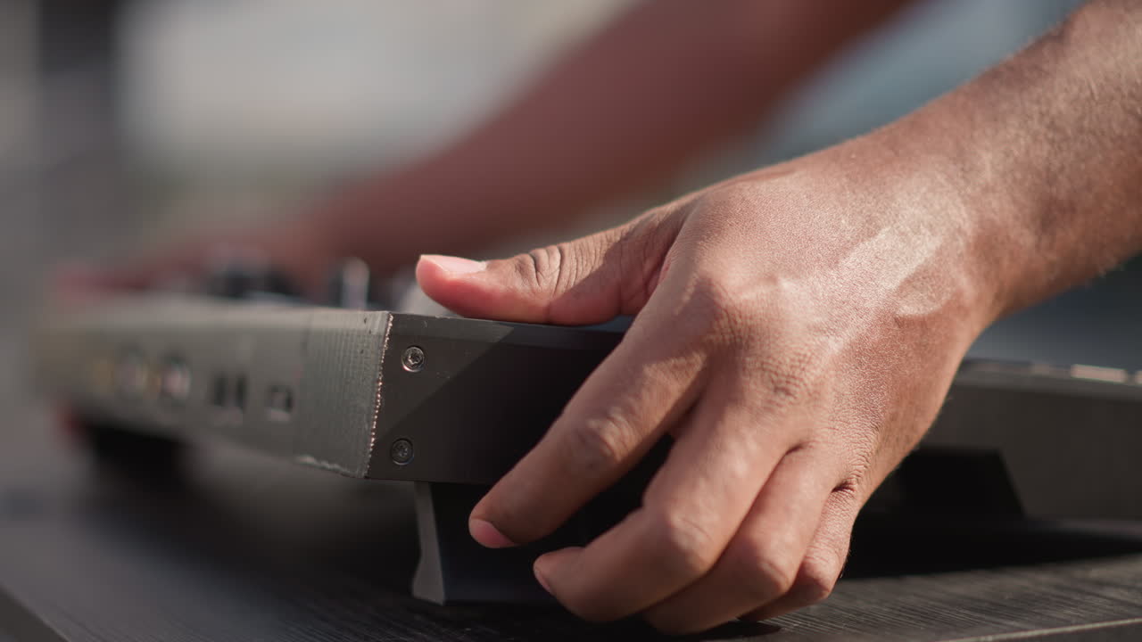 Mani in primo piano che caricano un controller DJ su un tavolo portatile sotto la luce del sole, manica di camicia rossa visibile, porte e piedini in gomma visibili, allineamento e connessione attenti, sottile atmosfera stradale, profondità di campo ridotta.
