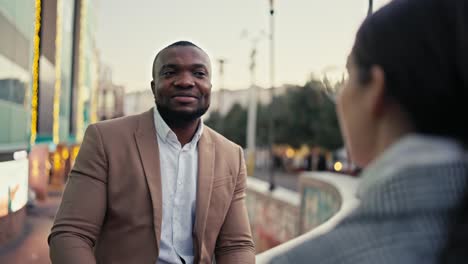over the shoulder a confident man with black skin with a short haircut and a beard in a brown jacket talks about his plans and decisions in the upcoming quarter during a business meeting with a businesswoman girl in the city
