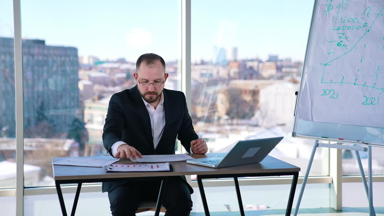 Busy man in suit working in office. Middle-aged entrepreneur in glasses sitting at table with many papers and a laptop and working seriously.