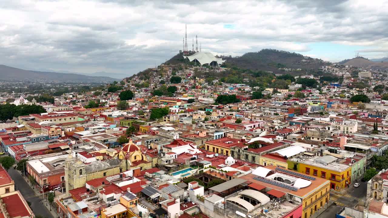 Drone push-in revealing the colorful urban fabric of Oaxaca City, moving toward Auditorio Guelaguetza as the focal point. Overcast daylight, soft contrast, mountains framing the skyline