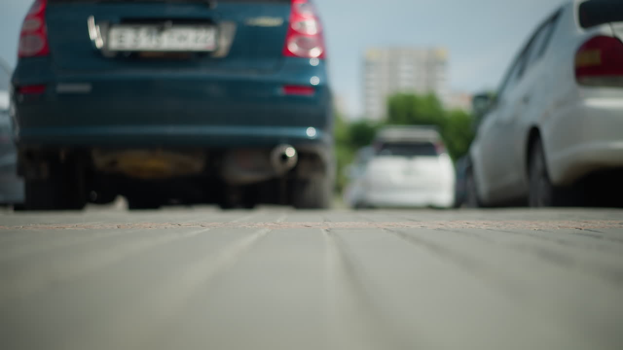 주차된 차량의 낮은 각도 시야에서 색 캔버스 스니커를 입은 사람이 지나가며, 땅에 그림자를 던지고, 먼 건물과 초록색이 흐릿한 배경에서 보입니다.