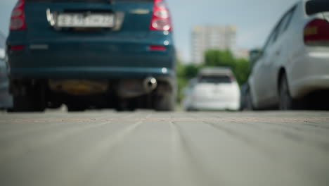 vista de ángulo bajo de coches estacionados con una persona en zapatillas de deporte de lona blanca pasando, proyectando sombra en el suelo, edificios distantes y vegetación visible en un fondo borroso