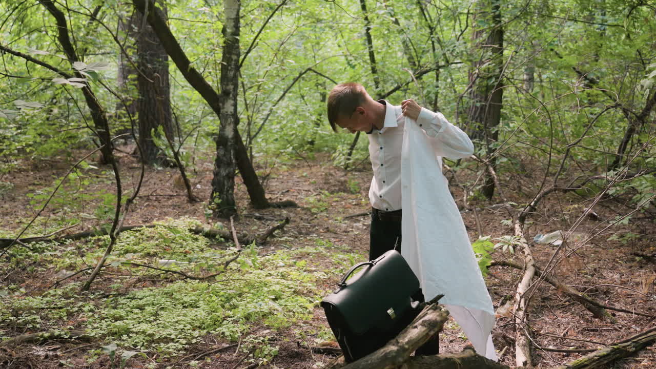 Young botany student in white shirt putting on white coat in forest near black leather bag resting on tree branch, surrounded by green leaves, trunks, branches, and natural sunlight