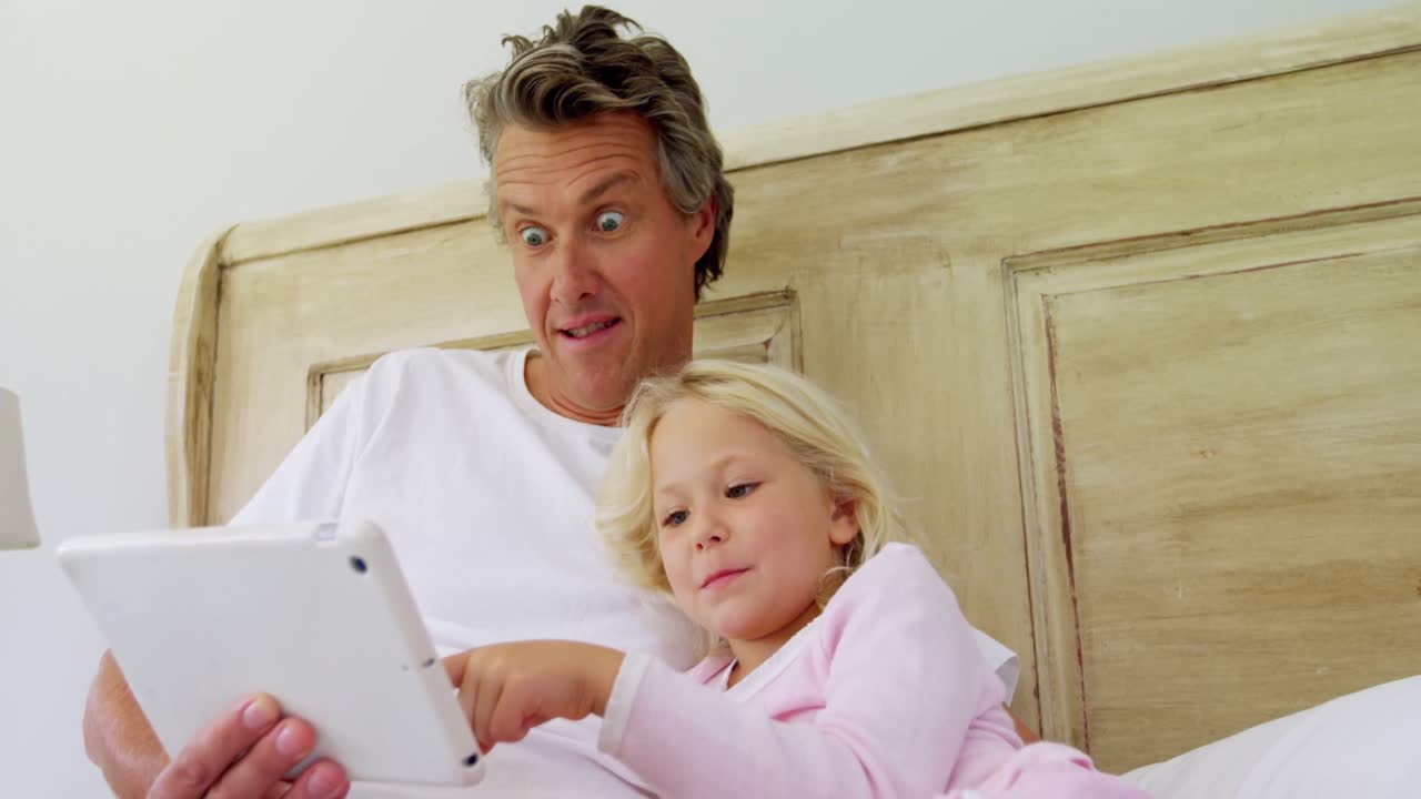 Smilende far og datter bruker nettbrett i seng 4k